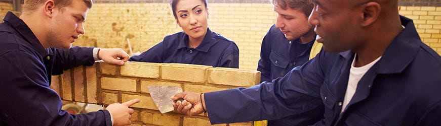Jeunes gens, en bleu de travail, se concertant autour d’un mur de briques en construction.