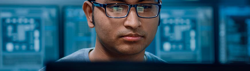 Jeune homme, de face, portant des lunettes, devant son ordinateur, travaillant sur des plans de circuits de câblages. En arrière-plan, d’autres écrans avec des schémas dans une ambiance de lumière bleue