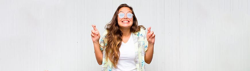 Jeune femme souriante, portant des fausses lunettes à filtre blanc et croisant les doigts en signe d’espoir.