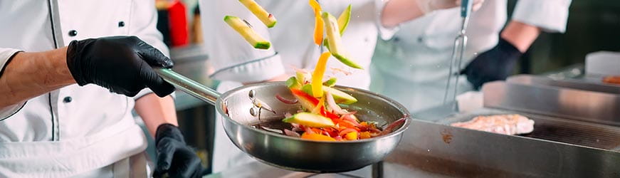  Plan serré de la main d’un cuisiner professionnel faisant sauter des légumes multicolores dans une poêle.