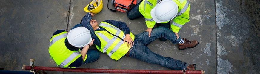 Scène d’accident sur un chantier. Deux personnes portent secours à un homme allongé à terre. Les trois individus portent un gilet jaune à bandes réfléchissantes.