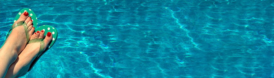 Pieds croisés dans des tongs vertes à pois, ongle vernis rouges. Le tout sur fond d’eau bleue de piscine.