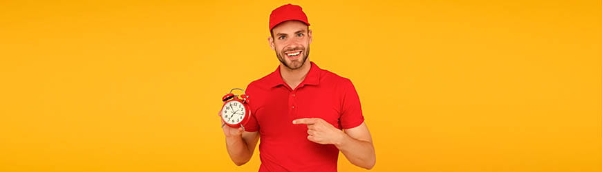 Jeune homme souriant sur fond uni jaune. Il porte un uniforme d’équiper, polo et casquette rouges. Il tient un réveil rouge qu’il pointe du doigt. 