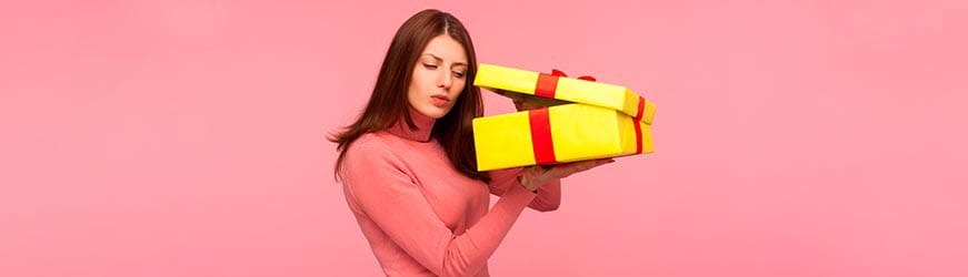 Jeune femme sur fond rose. Elle entrouvre un paque cadeau jaune à ruban rouge pour jeter un coup d’œil dedans.