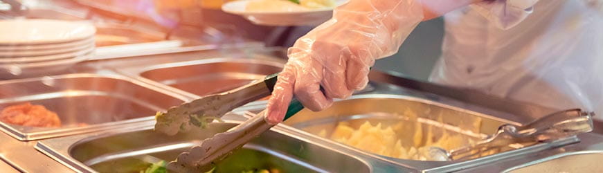 Gros plan sur une main servant des légumes verts dans une assiette. La personne au service porte des gants en latex et utilise une pince. La scène se passe dans un établissement type cantine.