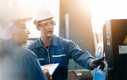 Deux hommes dans une usine, équipés de casques, lunettes de protection et blouses contrôlent le fonctionnement d'une machine