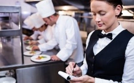 Femme cheffe dans un restaurant, dans ses cuisines, elle note sur un carnet une estimation de la main-d’œuvre dont elle aura besoin cet hiver 